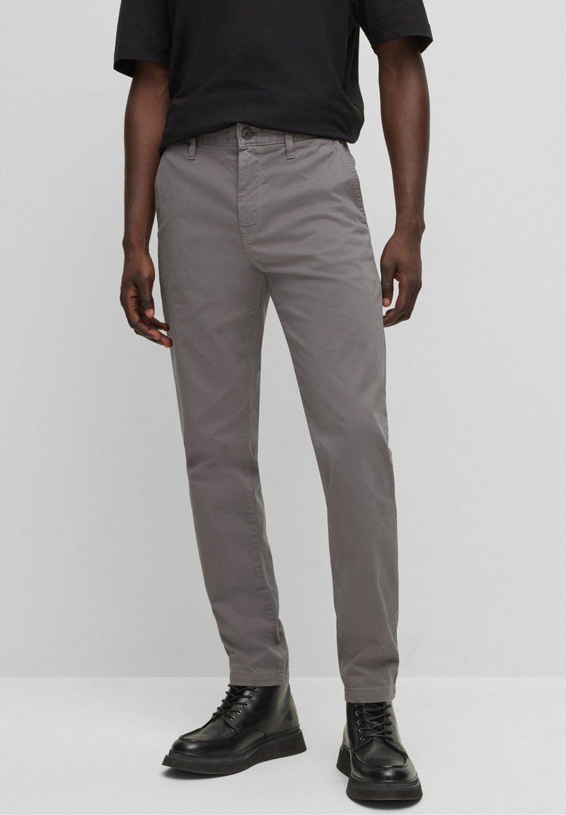 Homme portant un pantalon gris droit, des bottes noires à lacets et une chemise noire à manches courtes rentrée, debout devant un fond gris uni.