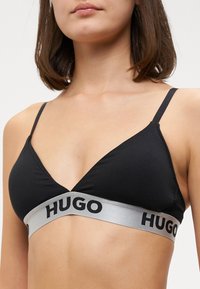 Γυναίκα που φοράει ένα μαύρο σουτιέν τριγωνικού σχήματος με λεπτές τιράντες και μια γκρίζα ελαστική ζώνη με την ένδειξη "HUGO" κάτω από το στήθος σε απλό φόντο.