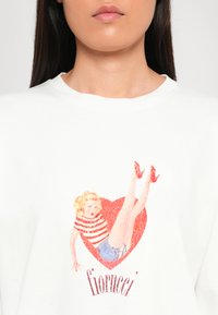 Hvit hettegenser med et grafisk motiv av en jente i et rødt hjerte, iført en stripete topp og blå shorts, med røde høyhælte sko og teksten 'fiorucci'.
