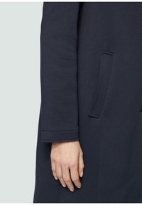 Cappotto blu navy con una texture di tessuto liscia, design semplice, tasche laterali e maniche sartoriali con orlo pulito.