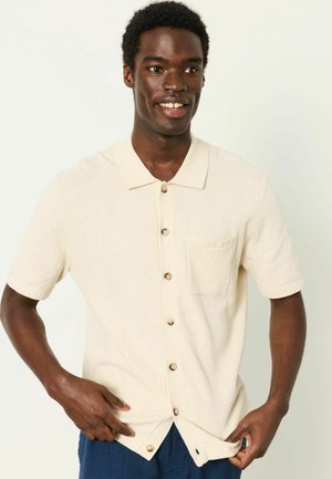 Lächelnder Mann in cremefarbenem Kurzarmhemd mit Knöpfen und Tasche, der den Saum anpasst und vor einem schlichten hellen Hintergrund steht.