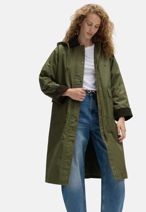 Vrouw draagt een lange olijfgroene mantel met capuchon over een wit shirt en blauwe spijkerbroek, met één hand die de voorkant van de mantel aanpast.