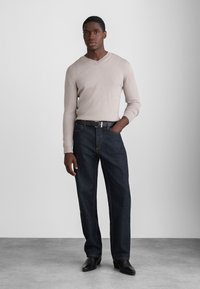 Bež pleteni pulover s V-izrezom, uparen s tamnoplavim ravnim trapericama. Outfit uključuje crni remen i crne kožne cipele.