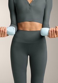 Žena drži svijetloplave bučice težine 1,5 kg, nosi tamno sive tajice viskog struka i skraćeni dugouhi top, tijelo i ruke su vidljivi.