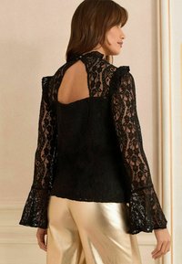 Blusa in pizzo nero con maniche lunghe e svasate e dettaglio schiena scoperta, caratterizzata da un colletto scallop e un motivo floreale. Abbinata a pantaloni dorati lucidi.