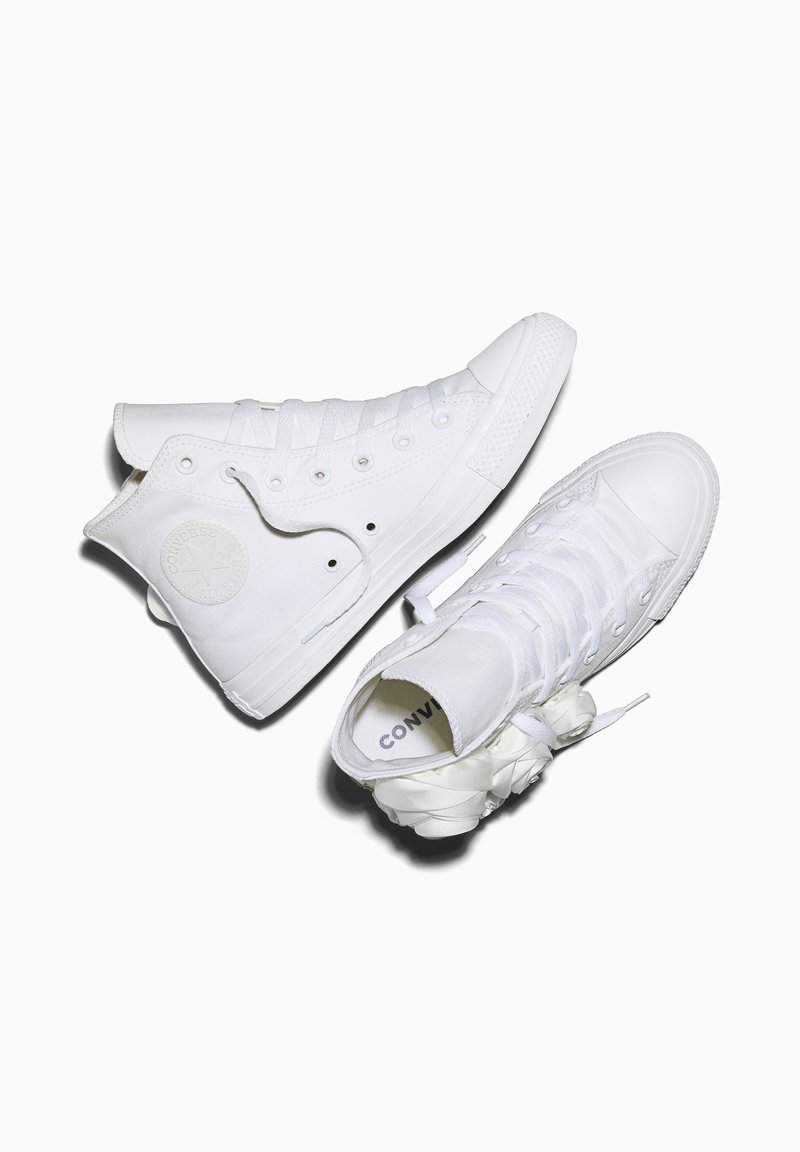 Een paar witte hoge Converse-sneakers met satijnen veterstrik, één schoen rechtop en de andere op zijn zijde op een witte achtergrond.