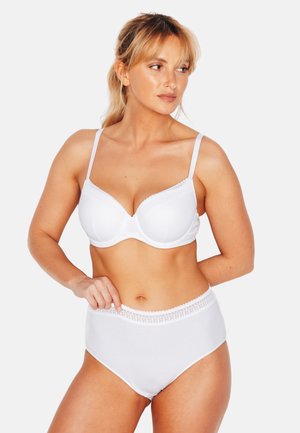 Donna dai capelli biondi che indossa un reggiseno bianco e mutandine a vita alta, in piedi con una mano sul fianco, guarda verso sinistra.