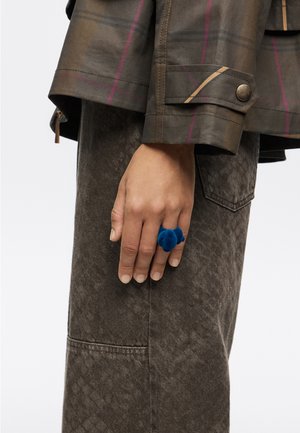 Anillo de terciopelo azul en una mano, combinado con un abrigo marrón texturizado que presenta patrones de cuadros y un puño abotonado.