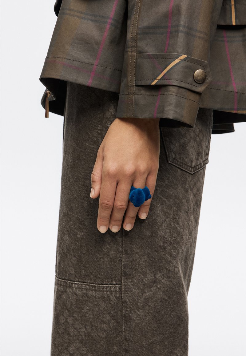 Zilgs samta gredzens uz rokas, kas savienots ar strukturētu brūnu mēteļi, kuram ir raibu rakstu un pogas aproces.