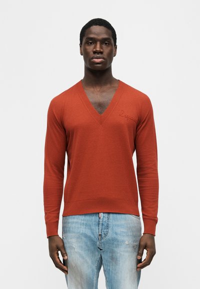 Homme portant un pull à col en V orange rouille et un jean bleu clair, debout devant un fond blanc uni, face à l'avant.