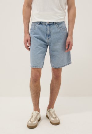 Hombre con pantalones cortos de mezclilla azul claro, camiseta blanca y zapatillas beige, de pie frente a un fondo liso.