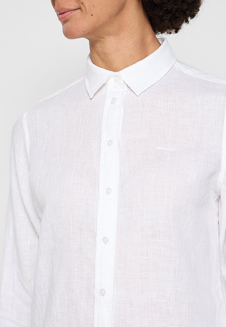 Biała lniana koszula z kołnierzykiem zapinanym na guziki, z przodem z plisą i subtelną teksturą. Małe logo po lewej stronie klatki piersiowej.