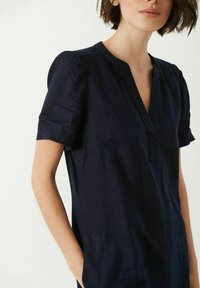 Navy blue linen blouse with short puff sleeves, a V-neckline, and a left side pocket; smooth texture with a relaxed fit.
