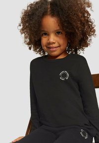 Kind met krullend haar, gekleed in een zwart shirt met lange mouwen en een broek met witte ronde tekstlogo's, zittend op een houten stoel voor een effen achtergrond.