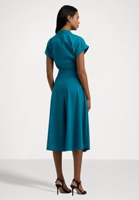 Vestido verde-água com mangas curtas, corpete ajustado e saia rodada. Tecido suave, até ao joelho com um detalhe de costura nas costas visível. Sapatos pretos de salto.