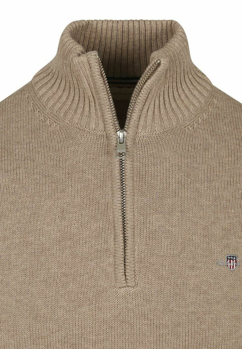 Beige zip-up tröja med ribbad krage och mudd, med en liten logopatch på den nedre sidan och ett texturerat stickat mönster.