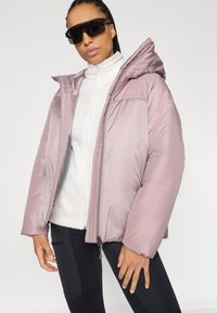 Giacca puffer lilla con zip frontale, cappuccio, maniche larghe e sottili motivi diagonali. Indossata sopra un top in pile bianco.