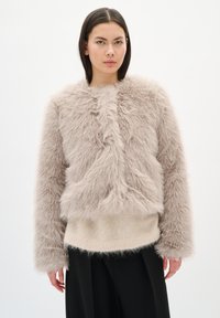 Chaqueta de piel sintética en beige claro con un exterior texturizado y esponjoso, cuello redondo, mangas largas y un corte cuadrado, llevada sobre un suéter beige.