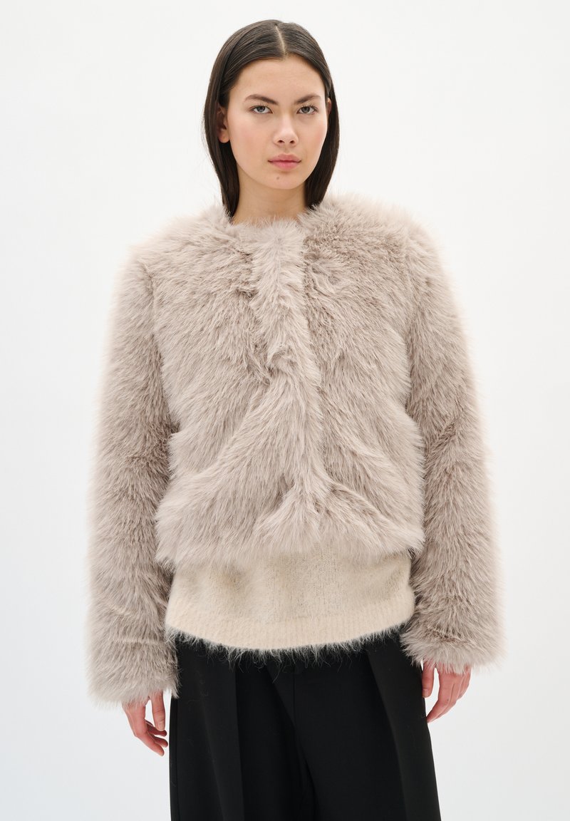 Chaqueta de piel sintética en beige claro con un exterior texturizado y esponjoso, cuello redondo, mangas largas y un corte cuadrado, llevada sobre un suéter beige.