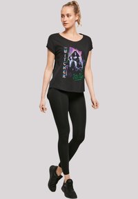 Black short-sleeve t-shirt featuring "The Witcher" graphic print in purple and green, paired with black leggings and black athletic shoes.