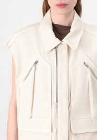 Gilet crema con colletto alto, chiusura con zip e grandi tasche frontali. Presenta accessori in tonalità argento e una finitura in tessuto testurizzato.