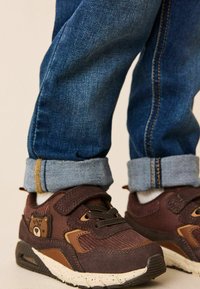 Brune og tan sneakers med velcro-rem og bjørnedetalje, parret med rullede denimjeans med en lyseblå manchet.
