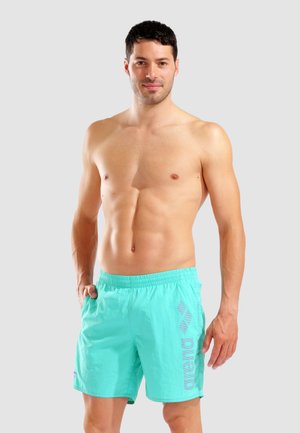 Uomo senza maglietta con capelli corti e scuri che indossa pantaloncini da bagno turchesi con il logo "arena", in piedi con una mano in tasca contro uno sfondo semplice.