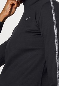 Czarna sportowa bluzka z długim rękawem i wysokim kołnierzem, z srebrnym paskiem po boku oraz małym logo na piersi. Gładka faktura.