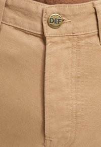 Pantalones de algodón beige con una superficie texturizada, cuentan con un botón dorado y una etiqueta marcada como "DEF" en la cintura.