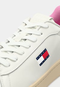 Vit Tommy Jeans-sneaker med vita snörningar, beige sula och röd innersulskant, med Tommy Jeans-logotypen på sidan.
