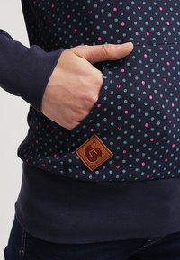 Mörkblå hoodie med teal- och rosa prickar, ribbade ärmslut och en framficka med en logotyp i brunt läder. Slät tygtextur.