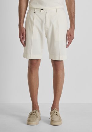 Hombre con pantalones cortos beige a medida y zapatos de cordones a juego sobre un fondo claro y liso.