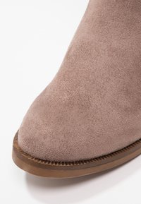 Bottine en daim taupe avec un bout arrondi et une surface texturée, dotée d'une semelle en caoutchouc marron contrastante avec des détails rainurés.
