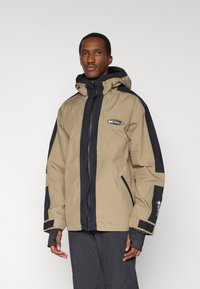 Αδιάβροχο μπουφάν σε μπεζ και ναυτικό μπλε με κουκούλα, φερμουάρ στο εμπρός μέρος, ελαστικές μανσέτες και λεπτομέρειες με λογότυπο. Διαθέτει δίχρωμο σχέδιο και ρυθμιζόμενη εφαρμογή.