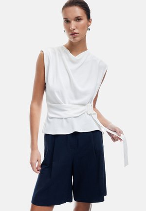 Haut sans manches blanc avec un col drapé, associé à un short marine foncé avec des plis. Le haut est ceinturé à la taille avec une sangle blanche.