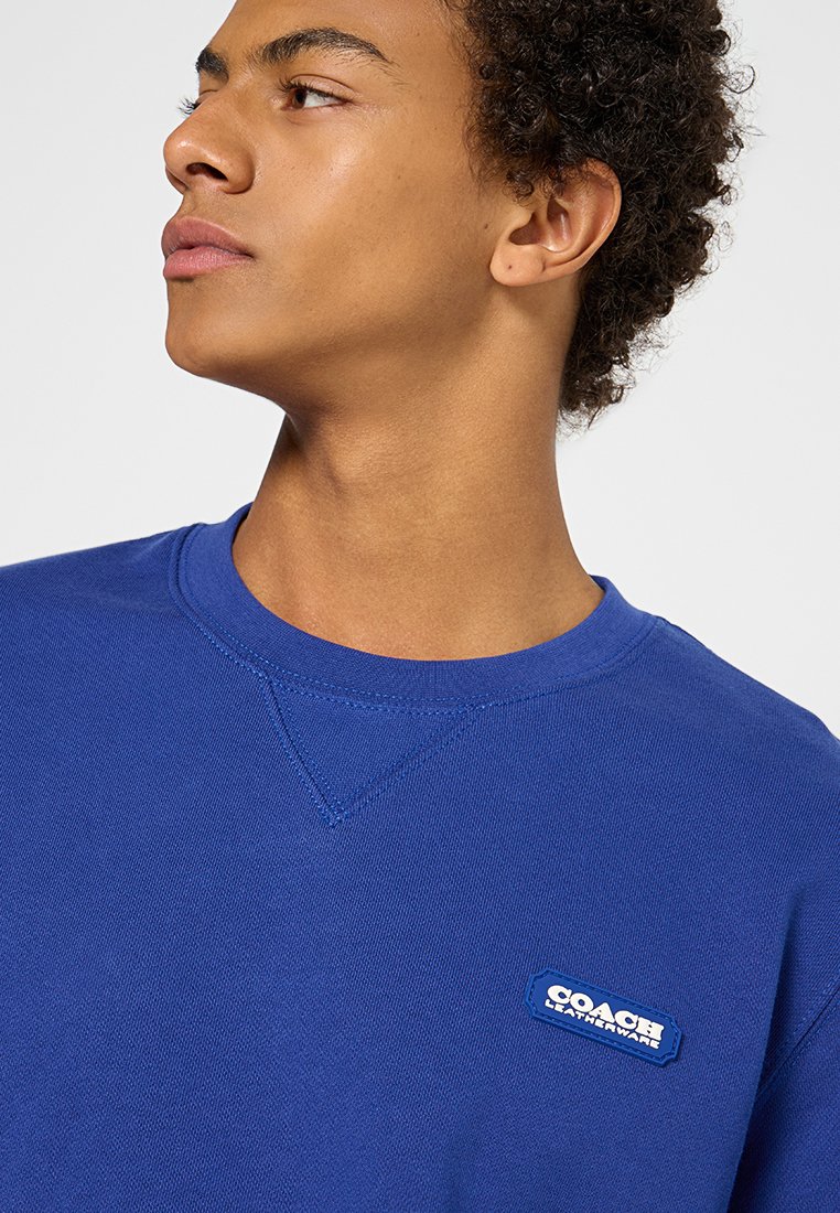 Sweatshirt bleu col rond avec une finition texturée et une petite étiquette blanche "COACH" sur la poitrine, présentant une coupe classique et un col côtelé.