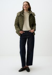 Olijfgroen pufferjack met een afneembare capuchon, geplooid patroon, geribbelde kraag, crèmekleurige trui en donkerblauwe wijde jeans. Zwarte instappers.