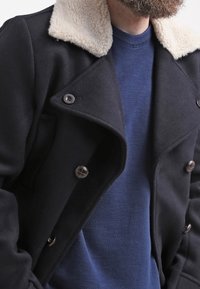 Svart dubbelknäppt kappa med en krämfärgad faux shearling-krage, stora mörka knappar och en mjuk textur, bärs över en blå tröja.