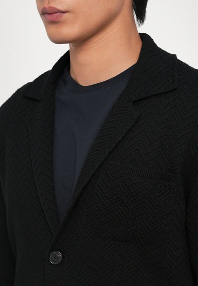 Melnā teksturētā austu žakete ar herringbone rakstu, ar pīķa atlokiem, vienas pogas aizdari un kabatu uz krūtīm, uz zila apaļā kakla krekla.