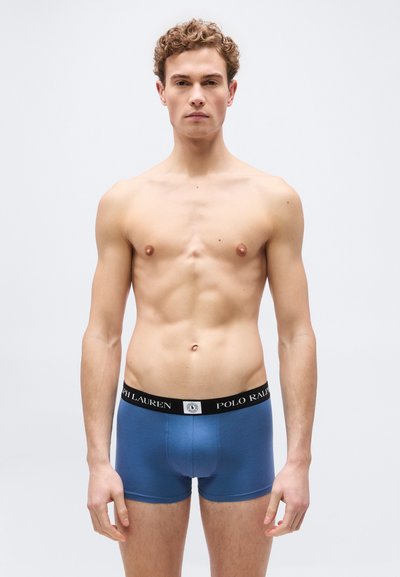 Νέος άνδρας με σγουρά μαλλιά που φοράει μπλε εσώρουχα Polo Ralph Lauren, στέκεται μπροστά σε ένα απλό ανοιχτόχρωμο φόντο.