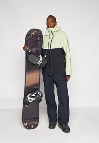 Snowboard con un motivo in legno scuro e attacchi in bianco e nero, tenuto da una persona che indossa una giacca leggera verde chiaro e nera, pantaloni blu navy e stivali neri.