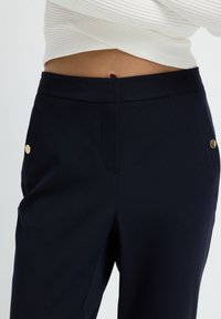 Close-up van de buik en marineblauwe broek met gouden knoopdetails op de zakken, gecombineerd met een witte geribbelde crop top.