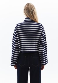 Marineblå och vit randig kort stickad tröja med breda ärmar, visad bakifrån, i kombination med högmidjade mörk denimjeans.