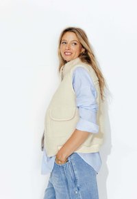 Gilet en polaire écru avec un col haut, deux poches avant, porté par-dessus une chemise rayée bleu clair et un jean léger.