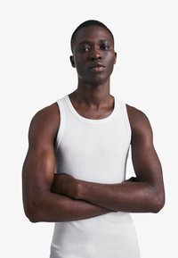 Mladi temnopolti moški s kratkimi lasmi, oblečen v belo majico brez rokavov, stoji z rokami prekrižanimi na preprostem belem ozadju.