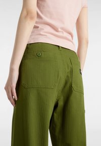 Gröna cargobyxor med texturerad tyg, med två bakfickor och en grön knapp. En ljusrosa t-shirt bärs ovanpå.