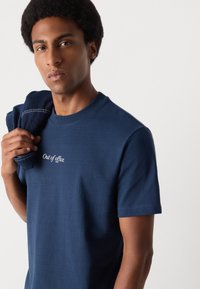 Camiseta de algodón azul marino con cuello redondo, con "Fuera de la oficina" bordado en blanco en la parte frontal. Textura suave, mangas cortas.