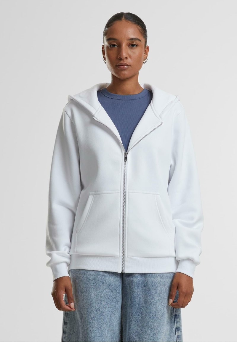 Felpa bianca con zip e texture morbida, caratterizzata da un ampio cappuccio, due tasche frontali e polsini a costine, indossata sopra una maglia blu navy.