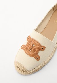 Kangas espadrille-kenkä vaaleassa kremissä, jossa on ruskeasta nahasta valmistettu logo-yksityiskohta, pyöreä kärki ja punottu jute-pohja, metallikoristeet ja ompeleet.
