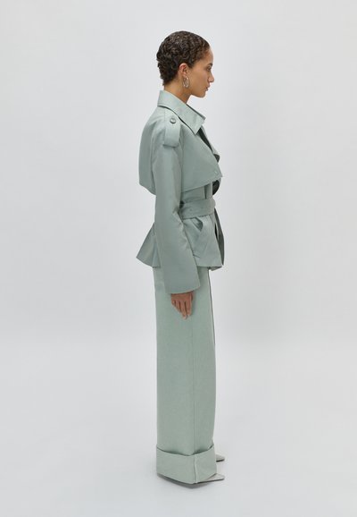 Γυναίκα με κοντά σγουρά μαλλιά που φοράει παλτό τραντσέρι ζαχαρί με ζώνη και παντελόνια με φαρδιά πόδια, στέκεται πλάγια μπροστά σε απλό φόντο.